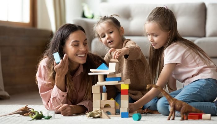 Happy mum with kids daughters playing toys on floor Happy young mum babysitter helping playing game with little kids daughters on floor, female nanny mother and cute small children sisters building castle of wooden blocks having fun with toys at home